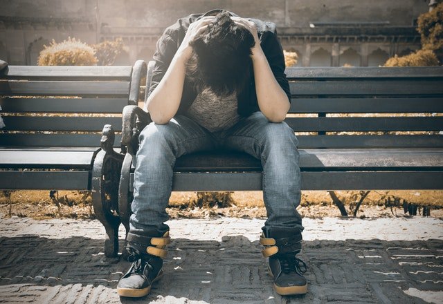 man sitting on bench with head tucked down staring at the ground