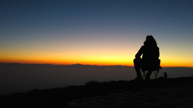 silhouette of a person sitting down in a field staring at a sunset sky